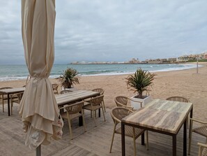 Ein Restaurant direkt am Strand mit Terrassensitzgelegenheiten, Blick auf das Meer und Gebäuden im Hintergrund.