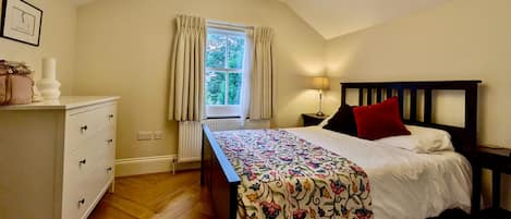 A bedroom with a bed, a bedside table, a lamp, a window with curtains, a radiator, and a dresser.