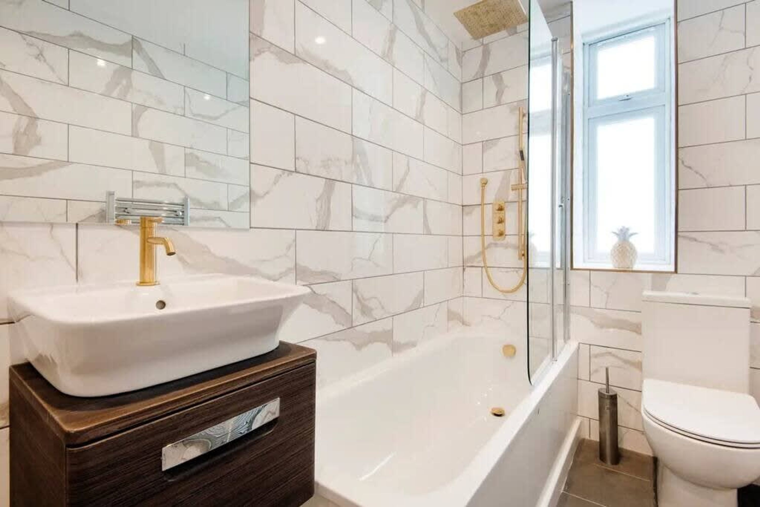 Et moderne badeværelse med væg af hvide marmorfliser, et hvidt badekar, en guldfarvet vandhane og et hvidt toilet.