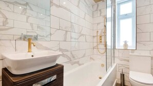 A modern bathroom with a white marble tiled wall, a white bathtub, a gold faucet, and a white toilet. - Stylish Ground-floor Retreat in Notting Hill (London)