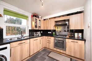 A modern kitchen with wooden cabinets, a black countertop, a built-in oven, and a window with a green blind. - Charming 1BR Flat in Sydenham With Free Parking (London)