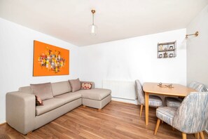 A modern living room with a beige sectional sofa, a wooden dining table with chairs, a white radiator, a wall-mounted shelf with decorative items, and a ceiling light fixture. - Charming 1BR Flat in Sydenham With Free Parking (London)