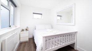 A white bedroom with a crib, a bedside table, a nightstand, a window, and a mirror on the wall. - Modern Apartment in Enfield With Comfort (Enfield)