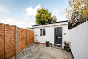 A small, single-story building with a black door and windows, surrounded by a wooden fence and a concrete ground. - Modern Apartment in Enfield With Comfort (Enfield)
