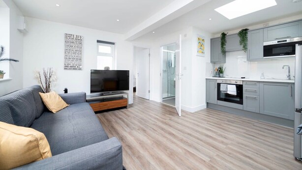 A modern apartment with a grey sofa, a flat-screen TV, a kitchen with grey cabinets, and a large window. - Modern Apartment in Enfield With Comfort (Enfield)