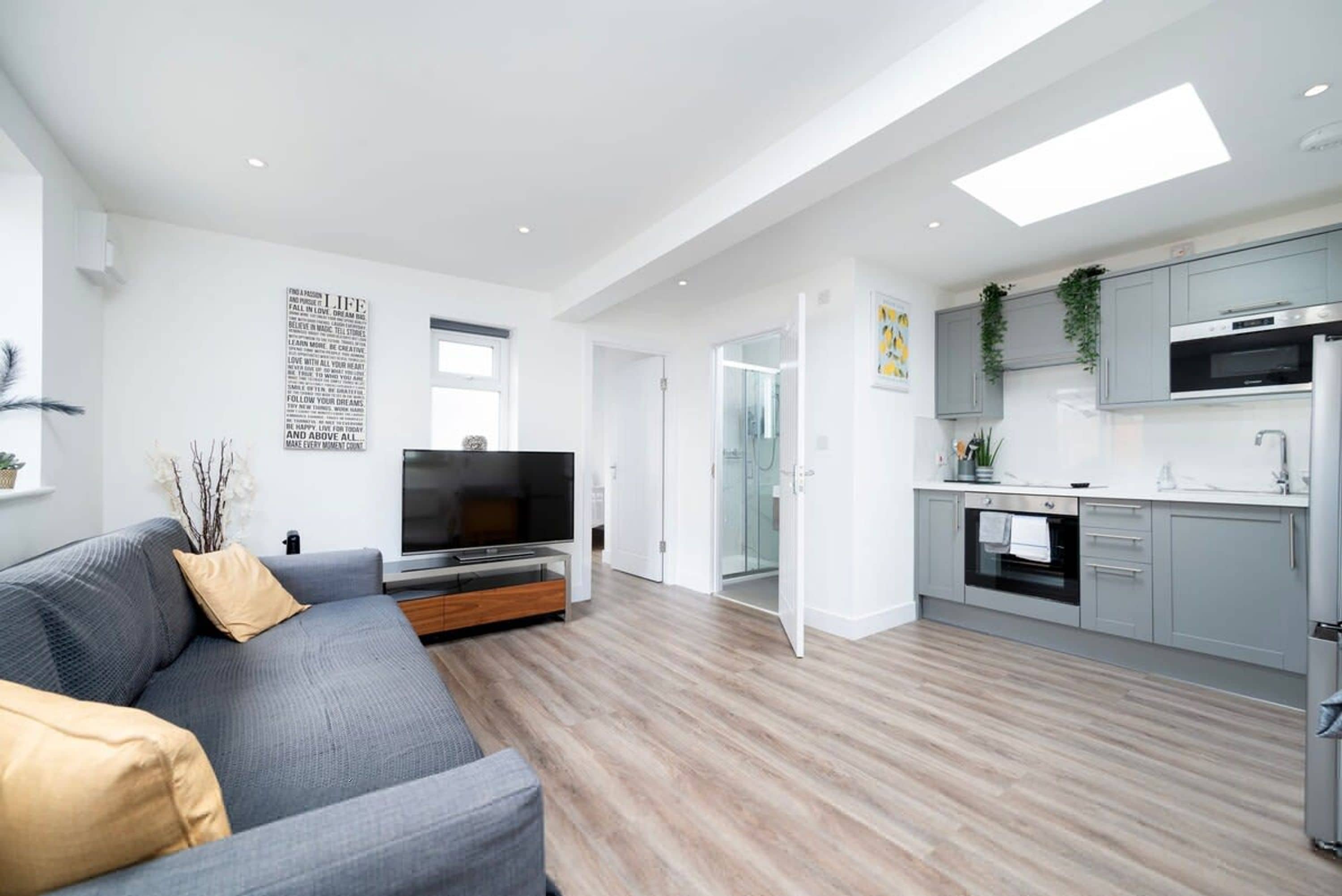 Eine moderne Wohnung mit grauem Sofa, einem Flachbildfernseher, einer Küche mit grauen Schränken und einem großen Fenster.