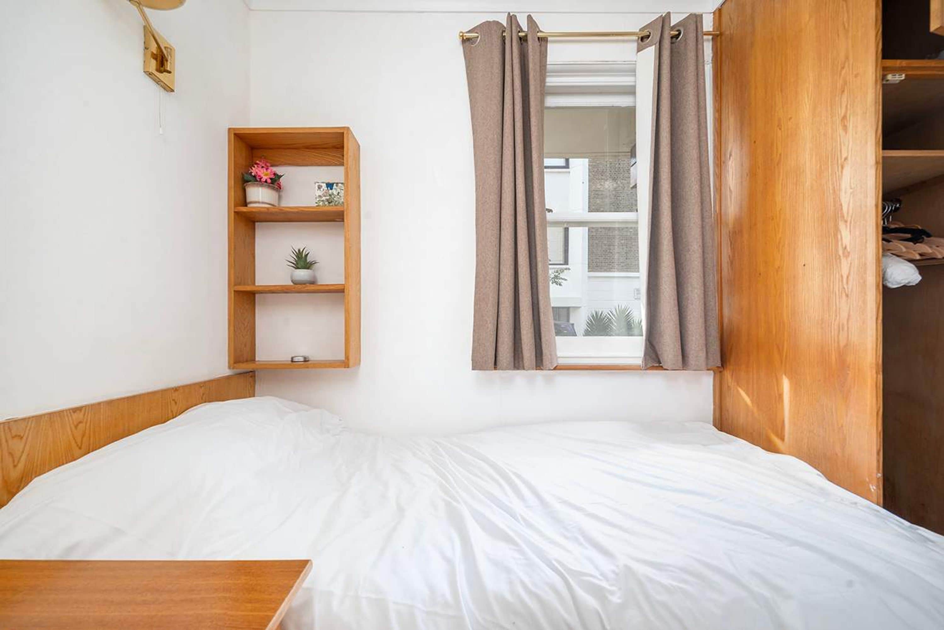 Ein Einzelbett mit Holzüberbau, eine kleine Holztrennwand mit Dekorationsobjekten und ein Fenster mit Vorhängen.