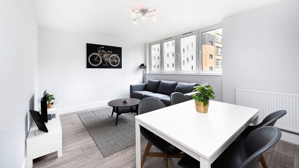 A modern, minimalist living room with a white sofa, a dining table, and a large window offering a view of buildings. - Charming 2-bedroom Apartment in London (London)