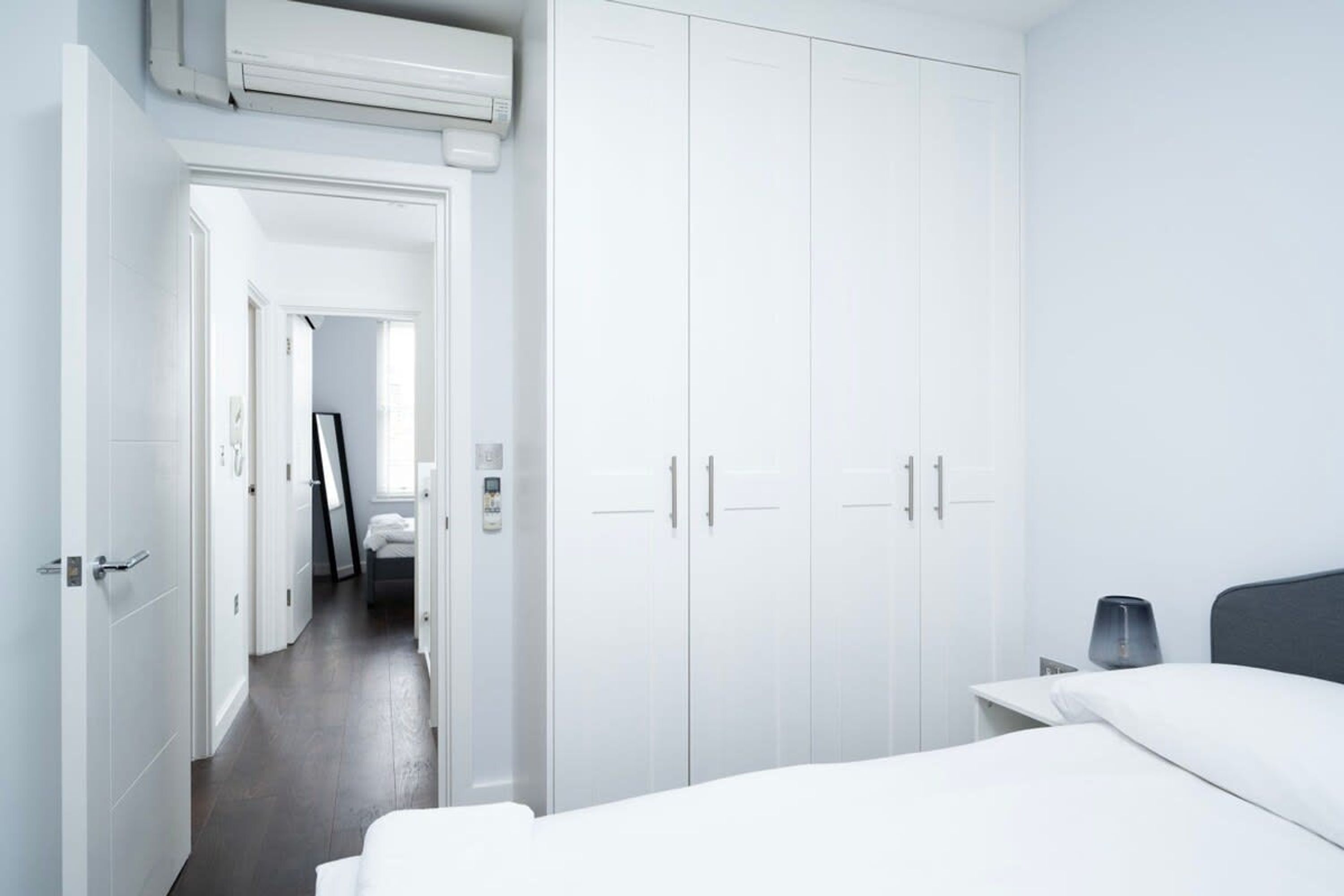 A modern hotel room with a large wardrobe, a bed, a bedside table, and a view of the hallway.