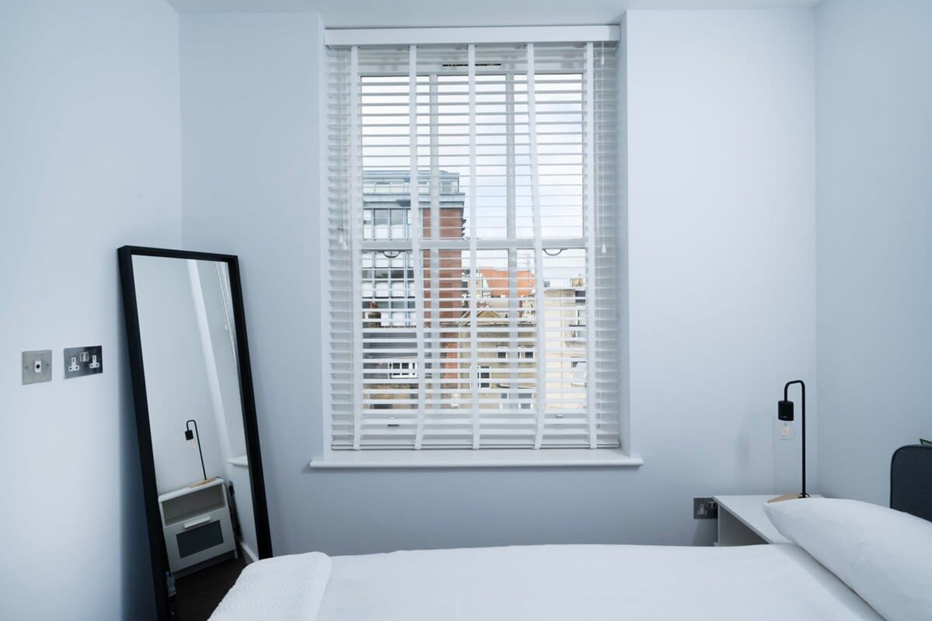 A modern hotel room with a bed, a bedside table, a lamp, a mirror, and a window with blinds.