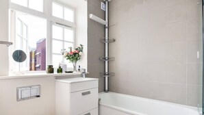 Een badkamer met een badkuip, toilet, raam met uitzicht op gebouwen en een plank met toiletartikelen.
