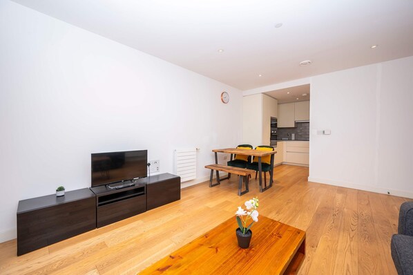 A modern living room with a flat-screen TV, dark wooden entertainment unit, wooden dining table with chairs, and a potted orchid on a wooden coffee table. - Modern Flat in Canning Town (London)