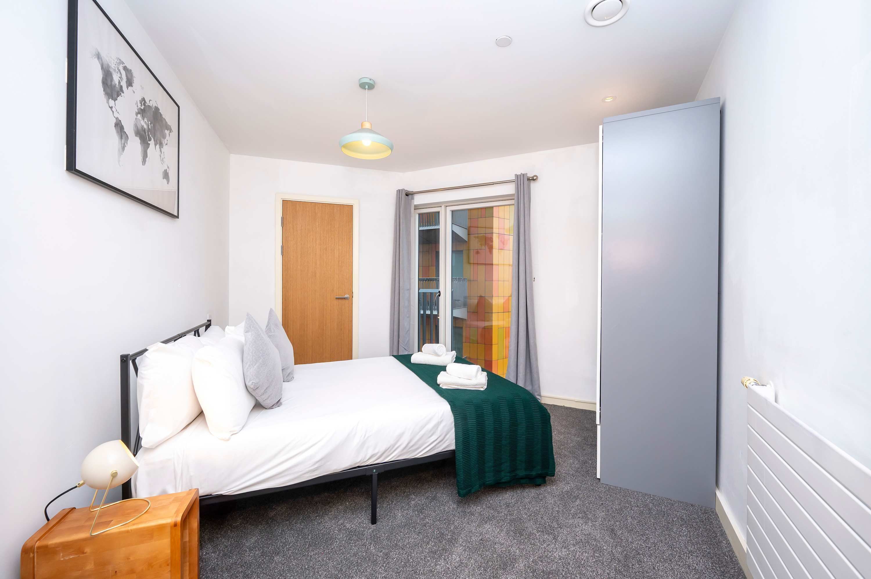 A hotel room with a bed, a bedside lamp, a framed world map, a window with curtains, and a radiator.