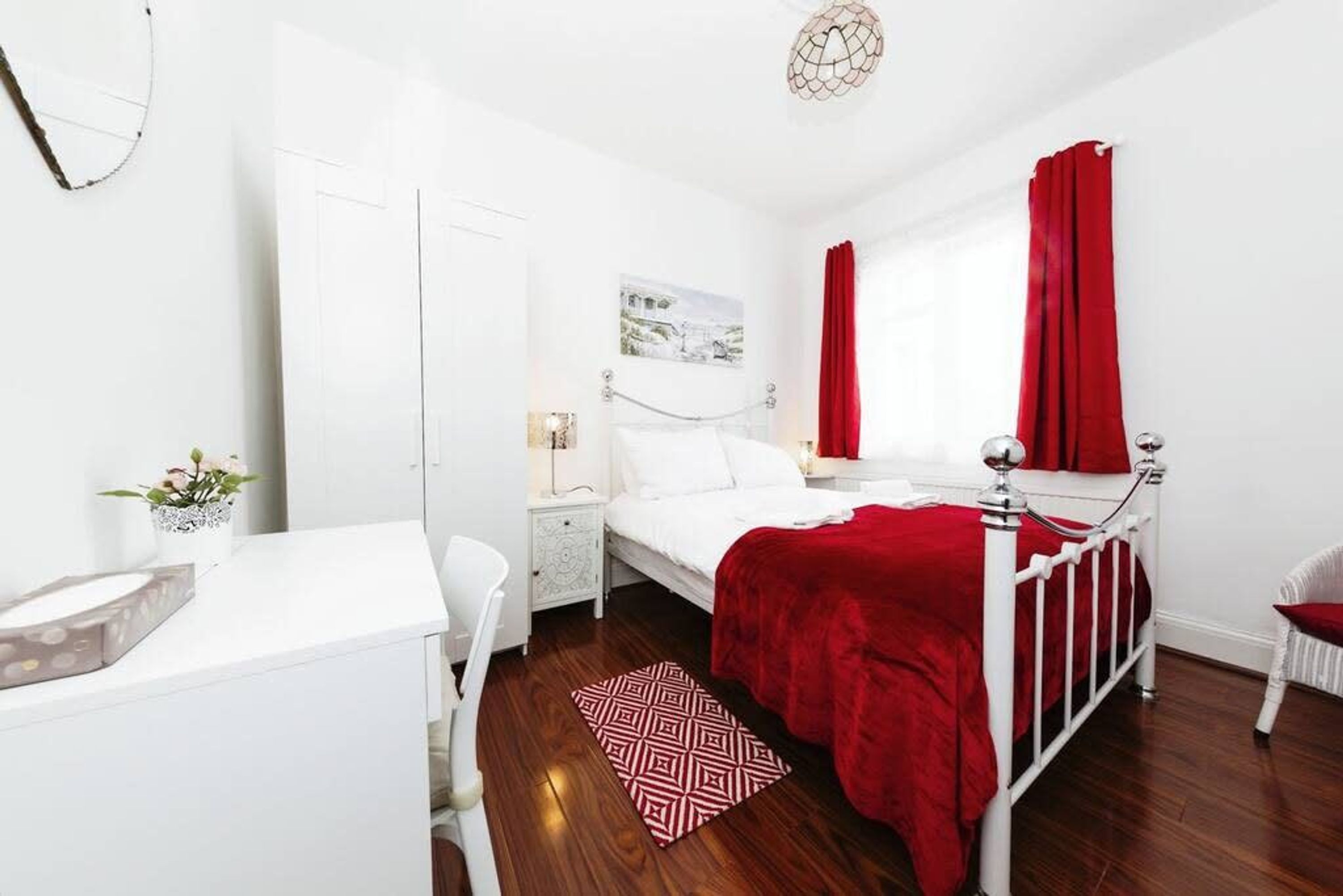 A bedroom with a white bed, red curtains, a white dresser, and a red and white patterned rug.