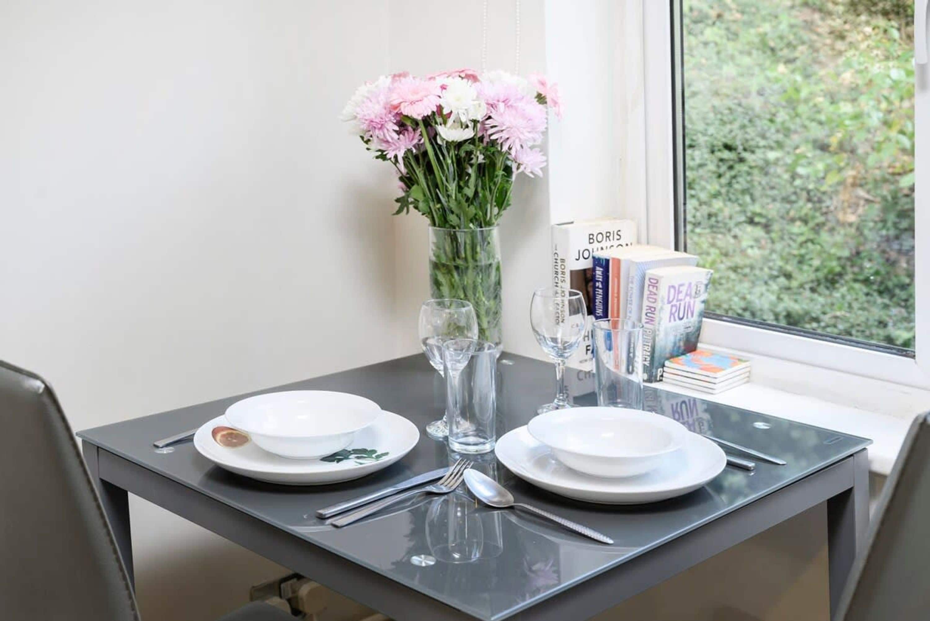 A dining table with plates, cutlery, a vase with flowers, and books by the window.