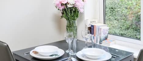 A dining table with plates, cutlery, a vase with flowers, and books by the window.