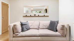 A neutral-toned sofa with two cushions, a wooden door, and a mirror above a shelf with small items. - Cozy One-bedroom Flat in Slough (Slough)