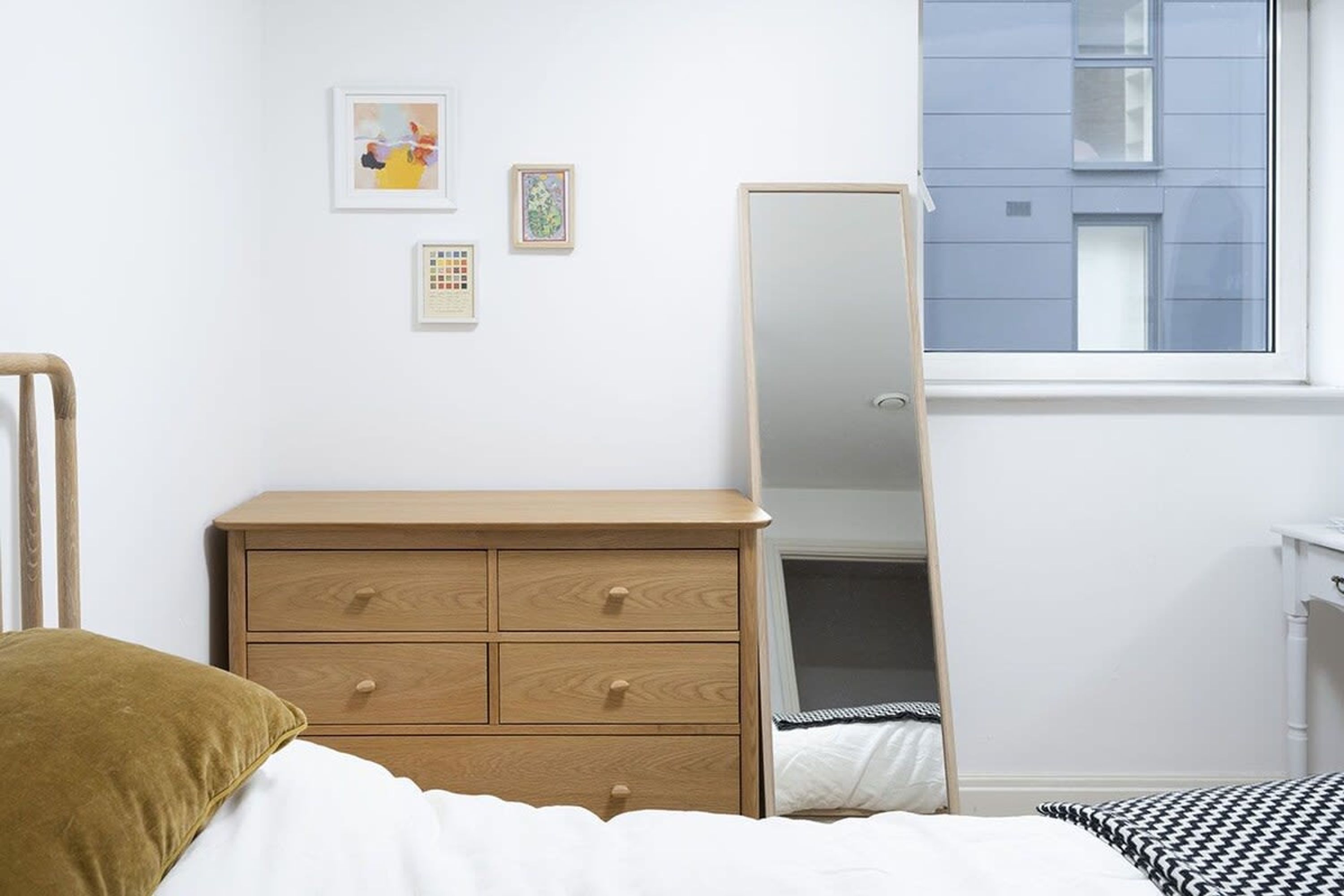 A bedroom with a bed, a wooden dresser, a mirror, and a window with a view of a building.