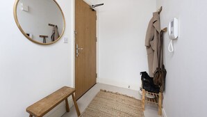 Um corredor com um banco de madeira, um espelho redondo, uma porta, um cabideiro com casacos e um sapateiro.