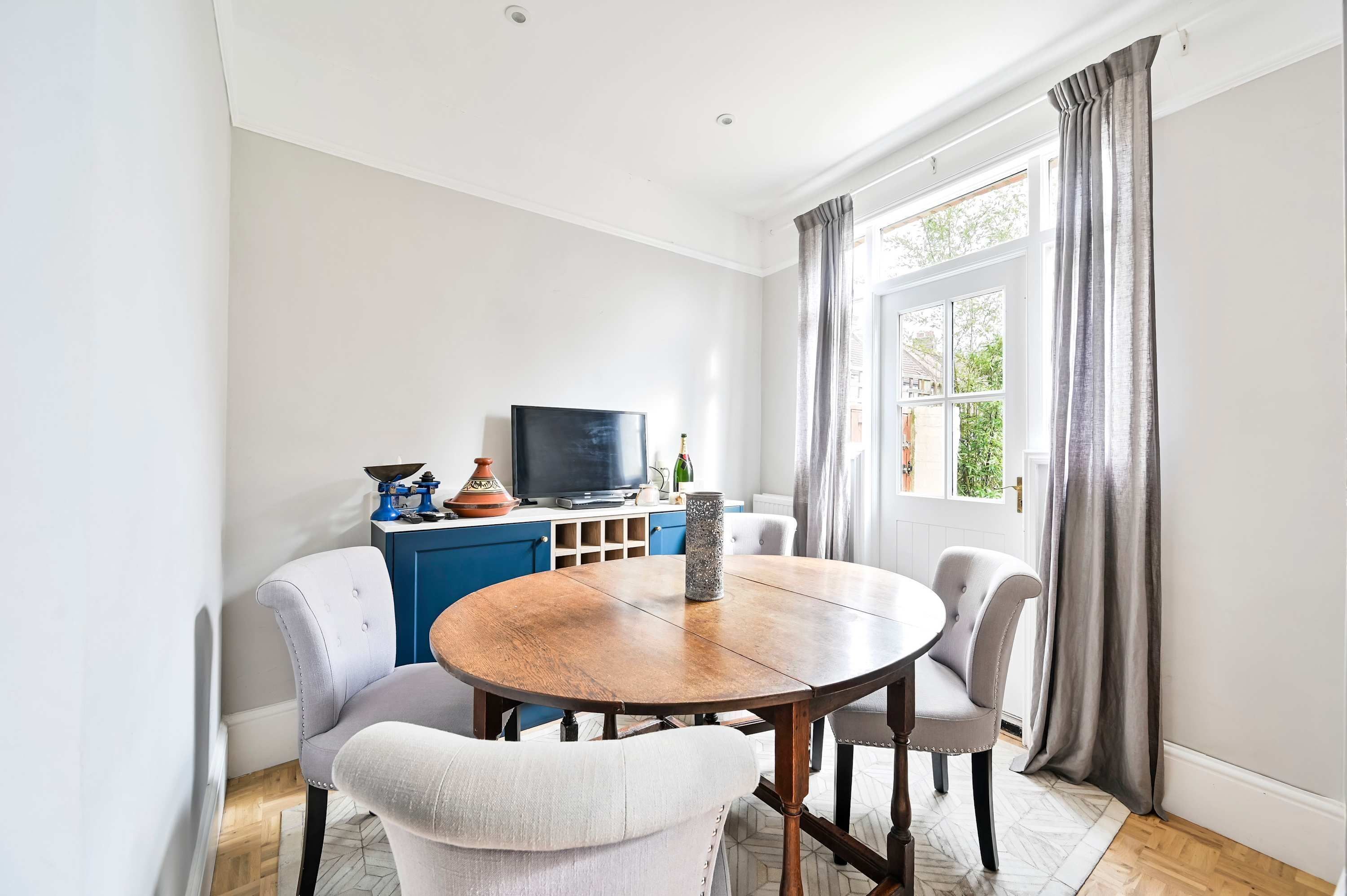 A dining area with a round wooden table, four upholstered chairs, a television on a blue cabinet, and a window with curtains.