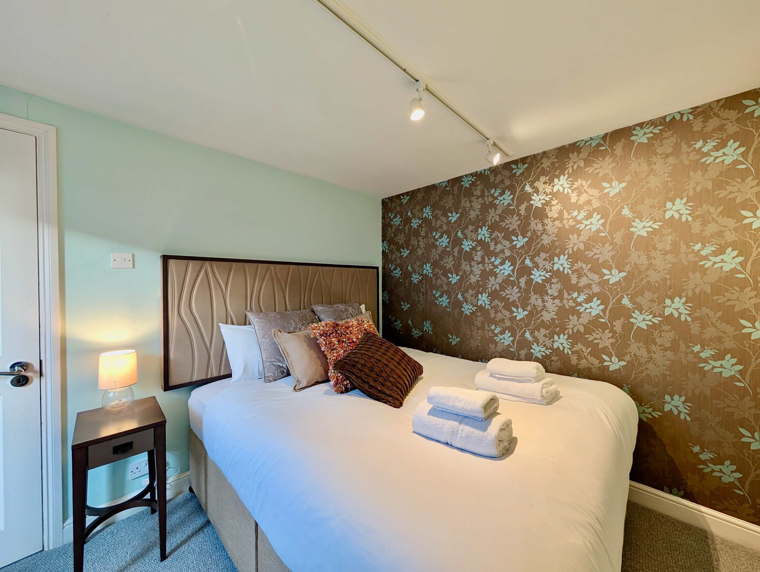 Een netjes opgemaakt bed met kussens en handdoeken, een nachtkastje met een lamp, en bloemenbehang aan de wand.