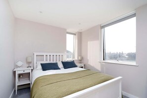 A bedroom with a white bed, two pillows, a green blanket, a bedside table with a lamp, and a window with a view. - Charming 1-bedroom Flat in London (London)
