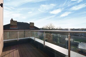 A balcony with a glass railing, wooden floor, and a view of houses and trees in the background. - Charming 1-bedroom Flat in London (London)