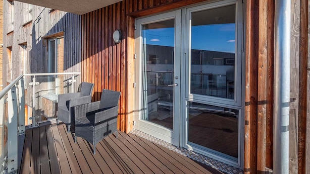 A wooden deck with outdoor furniture, a glass door leading to a covered area, and a metal railing. - Stunning 2BR Flat With Balcony (Maidenhead)