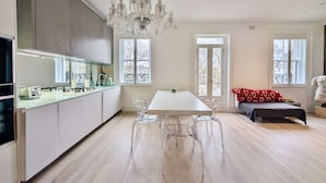 A modern kitchen with a central island, a dining area with a white table and clear chairs, and a chandelier. - Stylish 1 Bed in SW7 With Private Balcony (London)