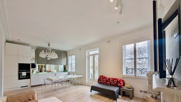 A modern living room with a sofa, a glass coffee table, and a bench. There is a kitchen area with a chandelier and a dining table. - Stylish 1 Bed in SW7 With Private Balcony (London)