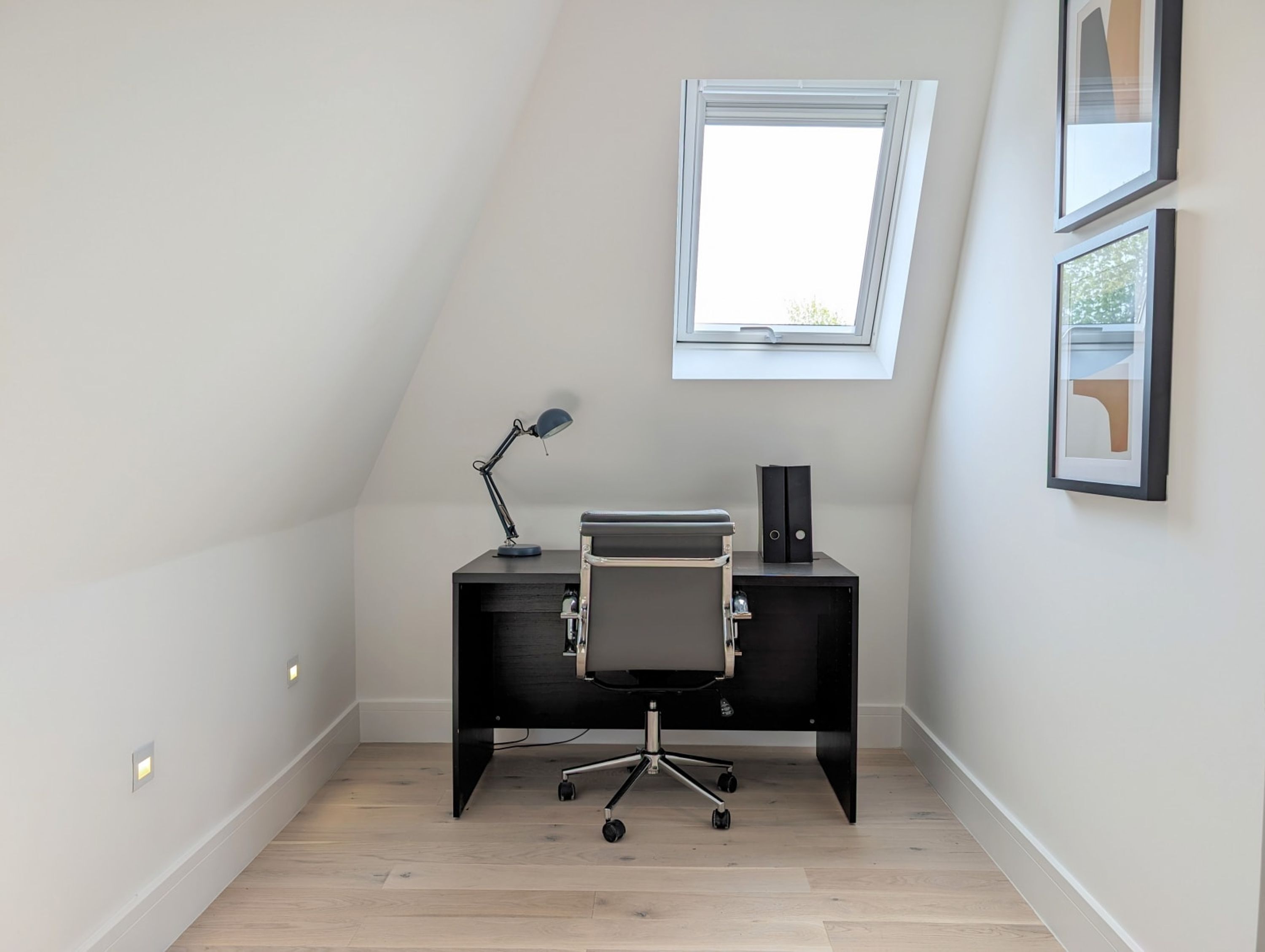 A small, modern office space with a desk, chair, and a single desk lamp. There is a window providing natural light and a framed picture on the wall.