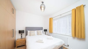 A neatly arranged bedroom with a bed, bedside tables, a wardrobe, and a window with curtains. - Charming 2-bedroom House in Northfleet (Gravesend)