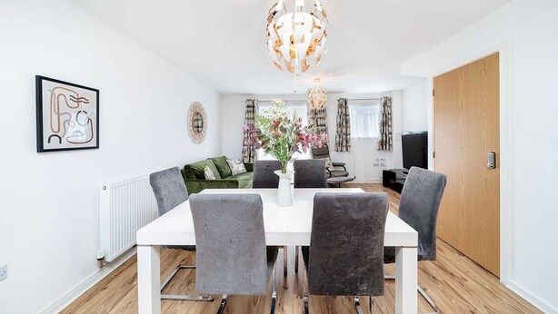 A modern dining area with a white table and grey chairs, a decorative chandelier, and a view into the living room. - Charming 2-bedroom House in Northfleet (Gravesend)