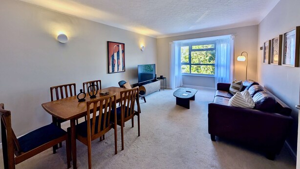 A living room with a dining table, chairs, a sofa, a TV, and a large window. - Charming Flat Near London Attractions (Surbiton)