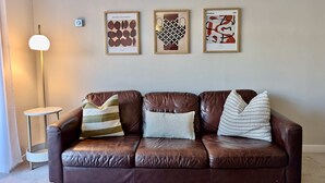 A brown leather sofa with striped and solid pillows, a floor lamp, and three framed artworks on the wall. - Charming Flat Near London Attractions (Surbiton)