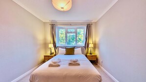 A neatly made bed with towels, a bedside lamp, and a window with curtains. - Charming Flat Near London Attractions (Surbiton)