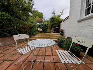 A patio with a round table and two chairs, surrounded by potted plants and a wooden staircase leading to a covered area. - Charming Two-bedroom House in London (Pinner)