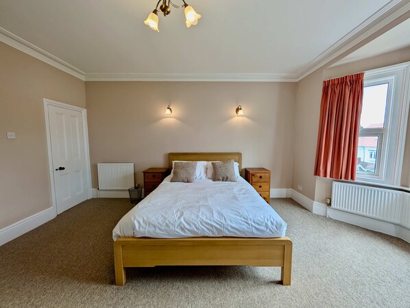 A bedroom with a wooden bed, two bedside tables, a radiator, a window with curtains, and a ceiling light fixture. - Charming Two-bedroom House in London (Pinner)