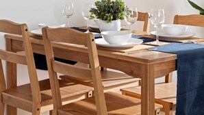 Un comedor con una mesa de madera preparada para dos personas, un reloj en la pared y una planta en maceta.