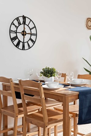 A dining area with a wooden table set for two, a wall-mounted clock, and a potted plant. - Brighton Getaway - Modern Apartment With Balcony (Brighton)