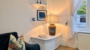 A modern living room with a dark blue sofa, a decorative pillow, a white side table with a lamp, a framed picture, and a window with sheer curtains. - Elegant 2-bed Flat in Shenfield (Brentwood)