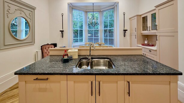A kitchen with a granite countertop, stainless steel sink, and light-colored cabinets. There is a window with blinds, a chandelier, and a decorative mirror on the wall. - Elegant 2-bed Flat in Shenfield (Brentwood)