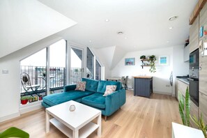 Ein modernes Wohnzimmer mit einem türkisfarbenen Sofa, einem weißen Couchtisch und einer Küchenzeile mit Holzschränken und einem Herd.