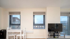 Une pièce moderne avec un bureau blanc, deux chaises, un téléviseur posé sur un meuble et de grandes fenêtres équipées de stores.