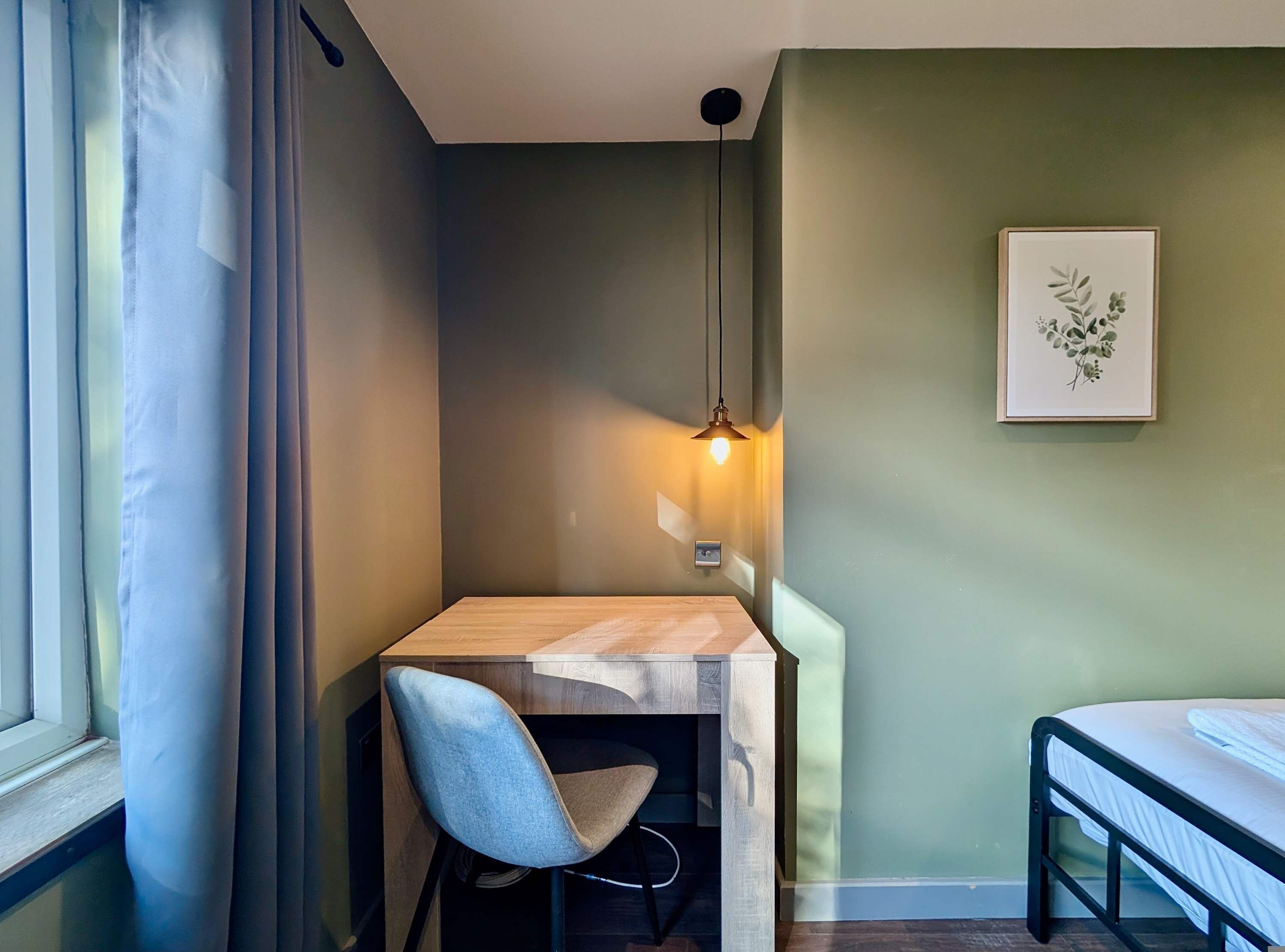 Een kamer met een houten bureau, een stoel, een bed, een wandlamp en een ingelijst schilderij.