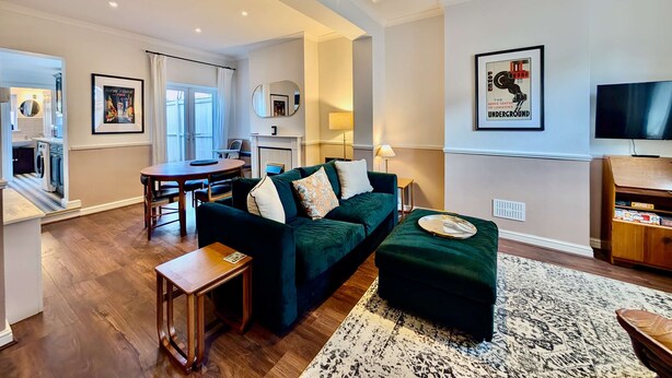 Un salon moderne avec un canapé vert, une table basse et un téléviseur. On y trouve également un coin repas avec une table et des chaises, et une cuisine visible au fond.