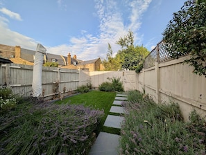 A backyard with a wooden fence, a paved pathway, and lavender plants. - Charming Two-bedroom Home in London (Sidcup)