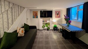 A modern living room with a dark grey sofa, green cushions, a flat-screen TV mounted on the wall, a bookshelf, a small table with a lamp, and a window with a view of the outside. - Charming House With Balcony Near Brighton Beach (Brighton)