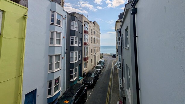 A narrow street lined with multi-story buildings, some with visible balconies, leading towards a body of water. - Charming House With Balcony Near Brighton Beach (Brighton)