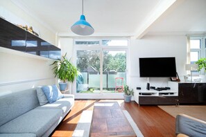 A modern living room with a grey sofa, a flat-screen TV mounted on the wall, a blue pendant light, and a large sliding glass door leading to a backyard.